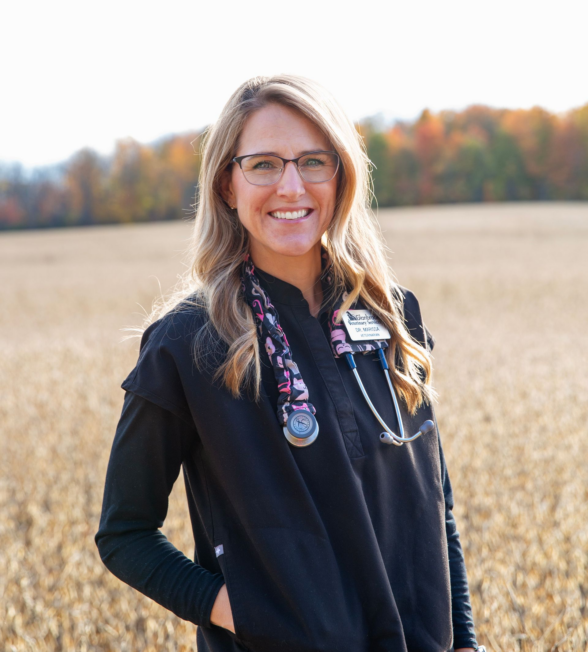Woman with arms crossed, smiling in a field; wearing a black shirt and red headband; autumn foliage in the background.