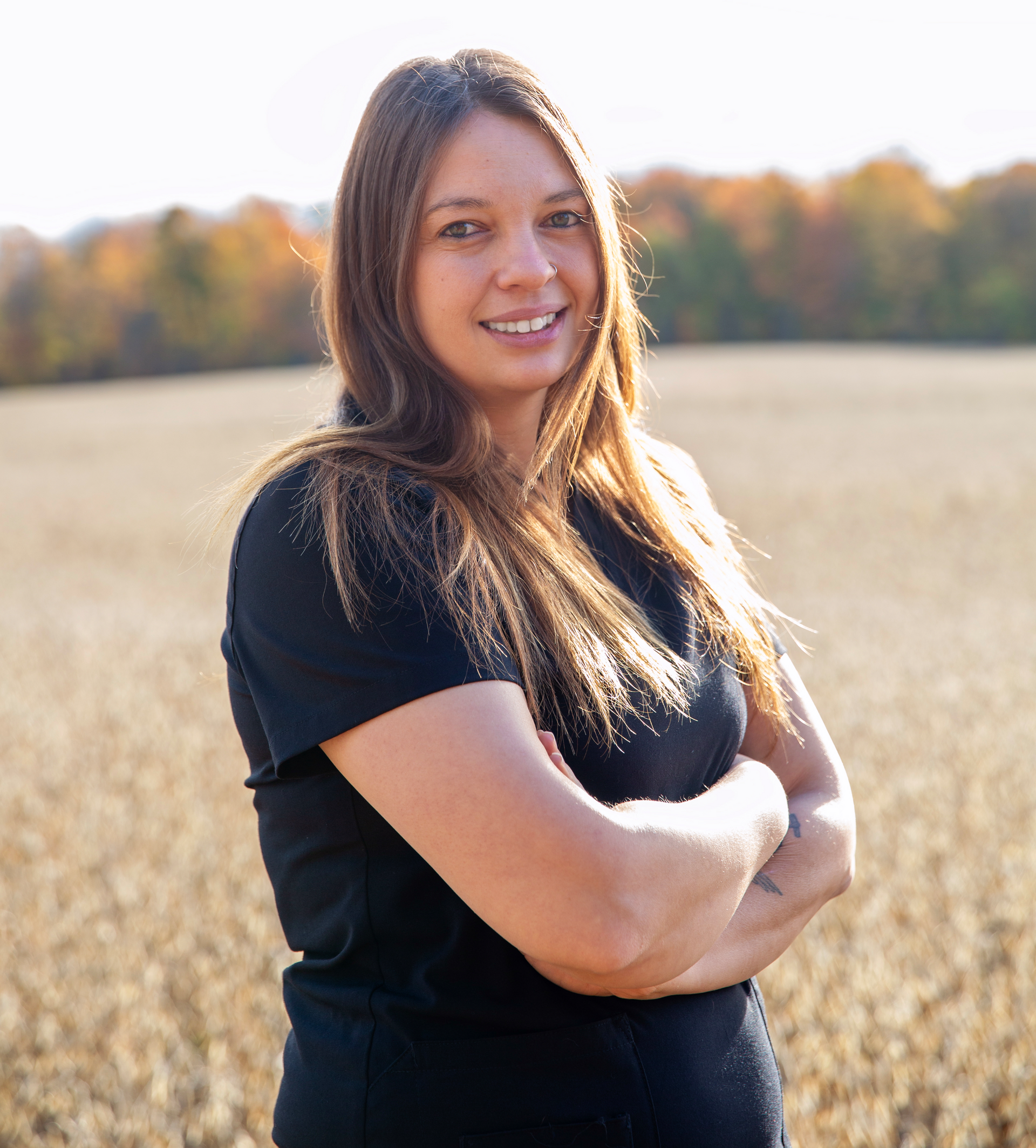Woman with crossed arms smiles in a field, trees in the background, daytime.