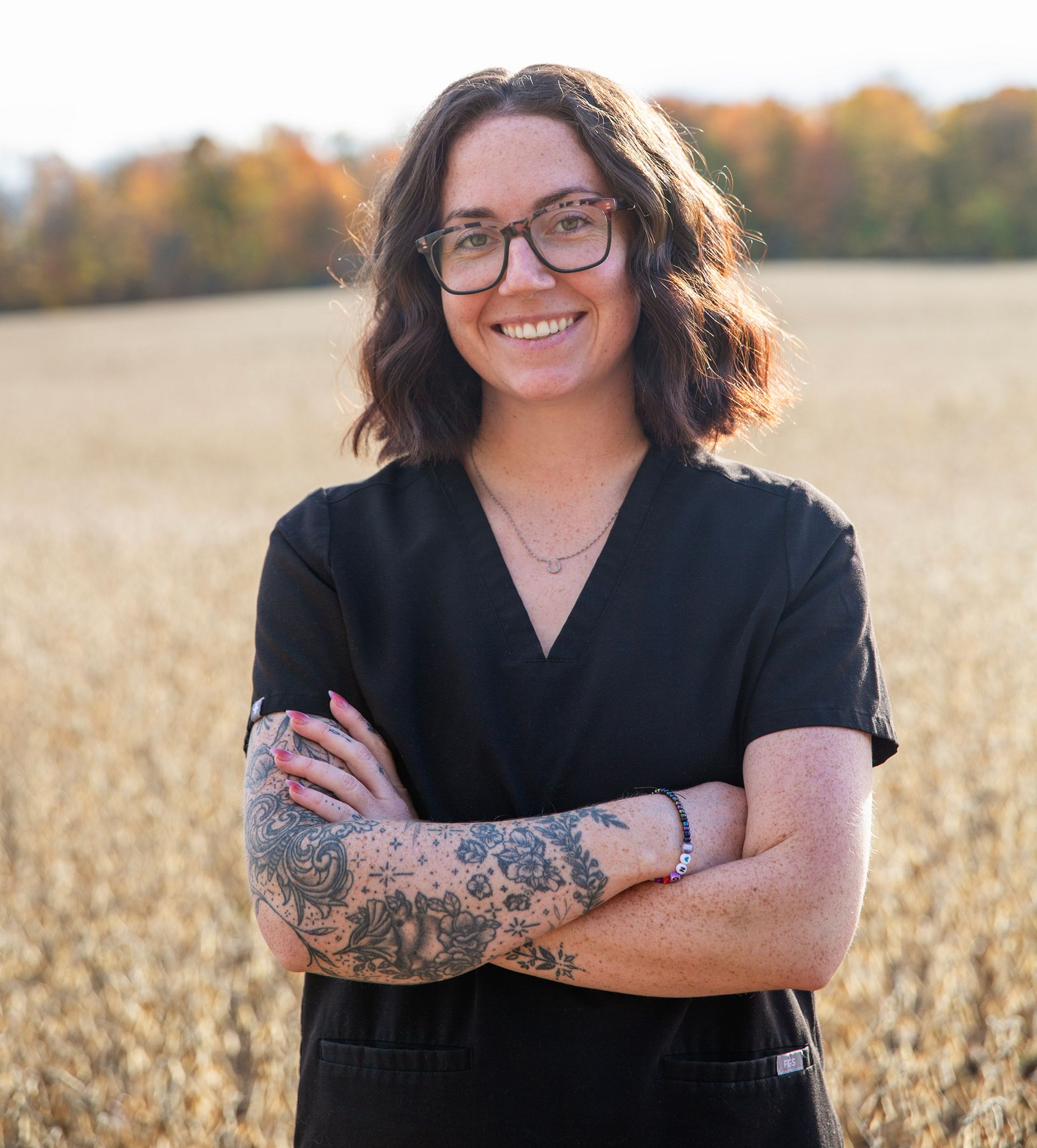 Woman in black scrubs with crossed arms, smiling outdoors, tattoos on arm, field background.