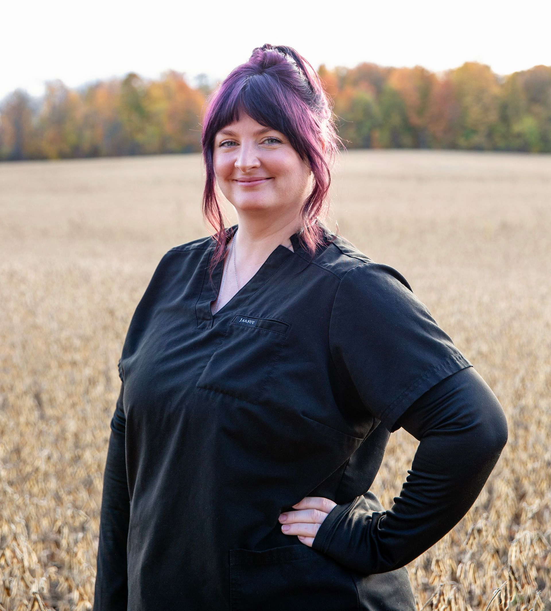 Woman in black scrubs stands in a field, hand on hip, purple-streaked hair, smiling.