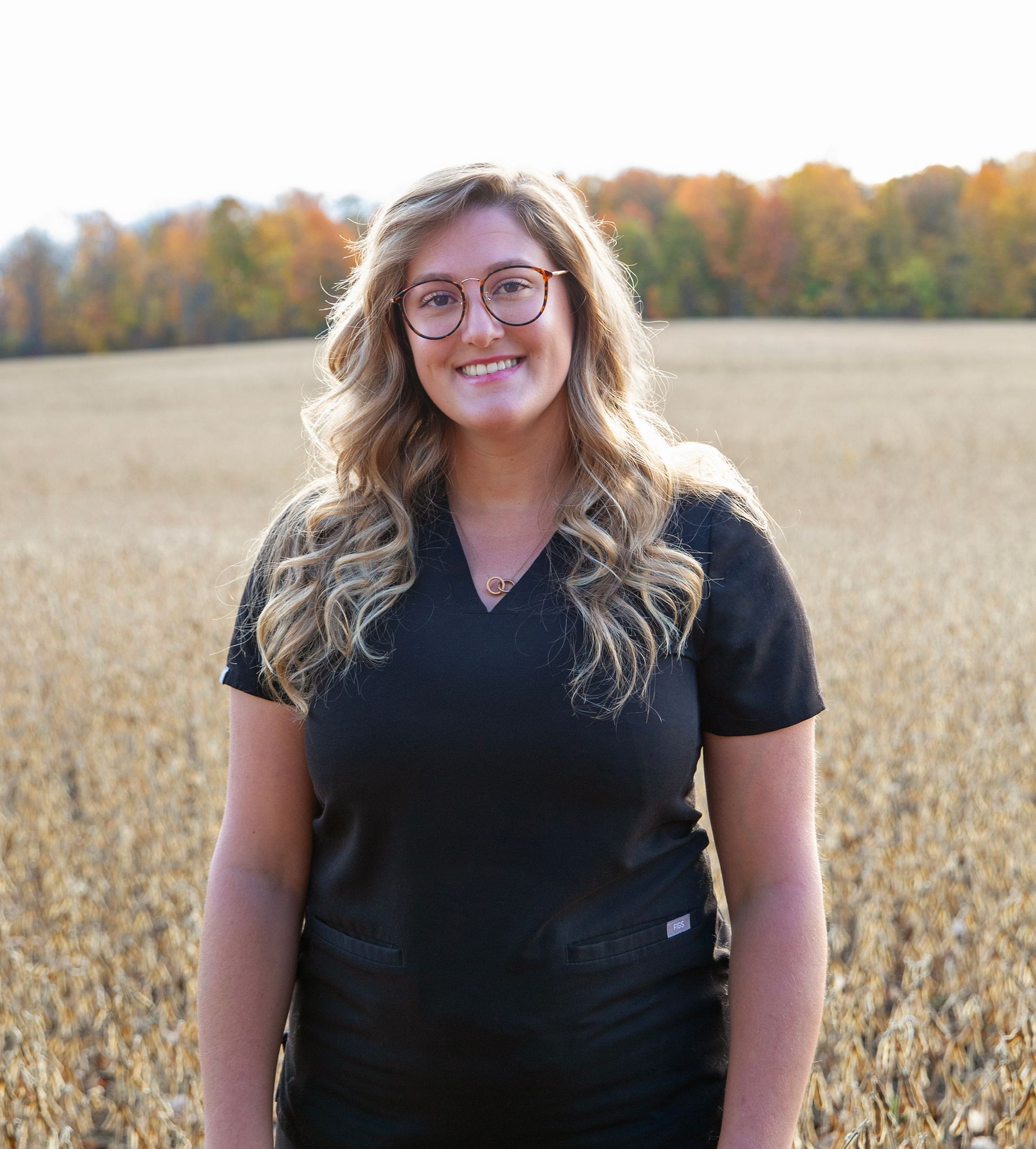 Woman smiling in black scrubs, glasses, field background.