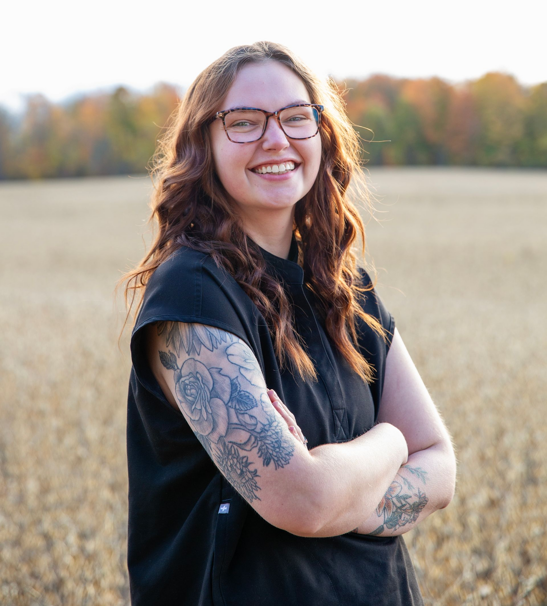 Woman with glasses smiles, arms crossed, stands in a field. Tattoo on arm, wearing black, with autumn trees in background.