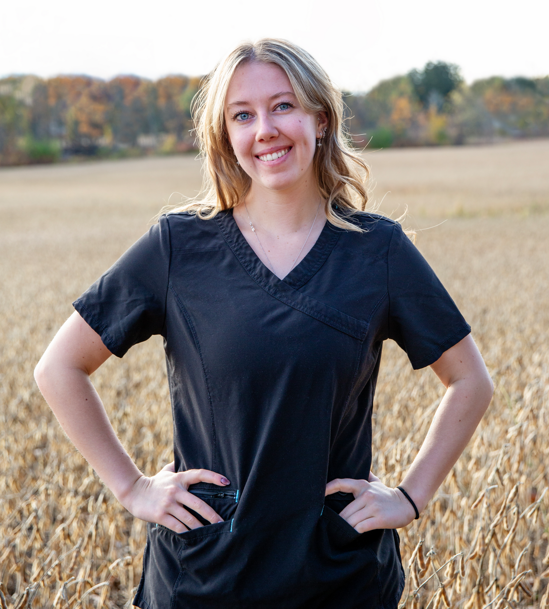 Woman in black scrubs, hands on hips, smiles in a field.