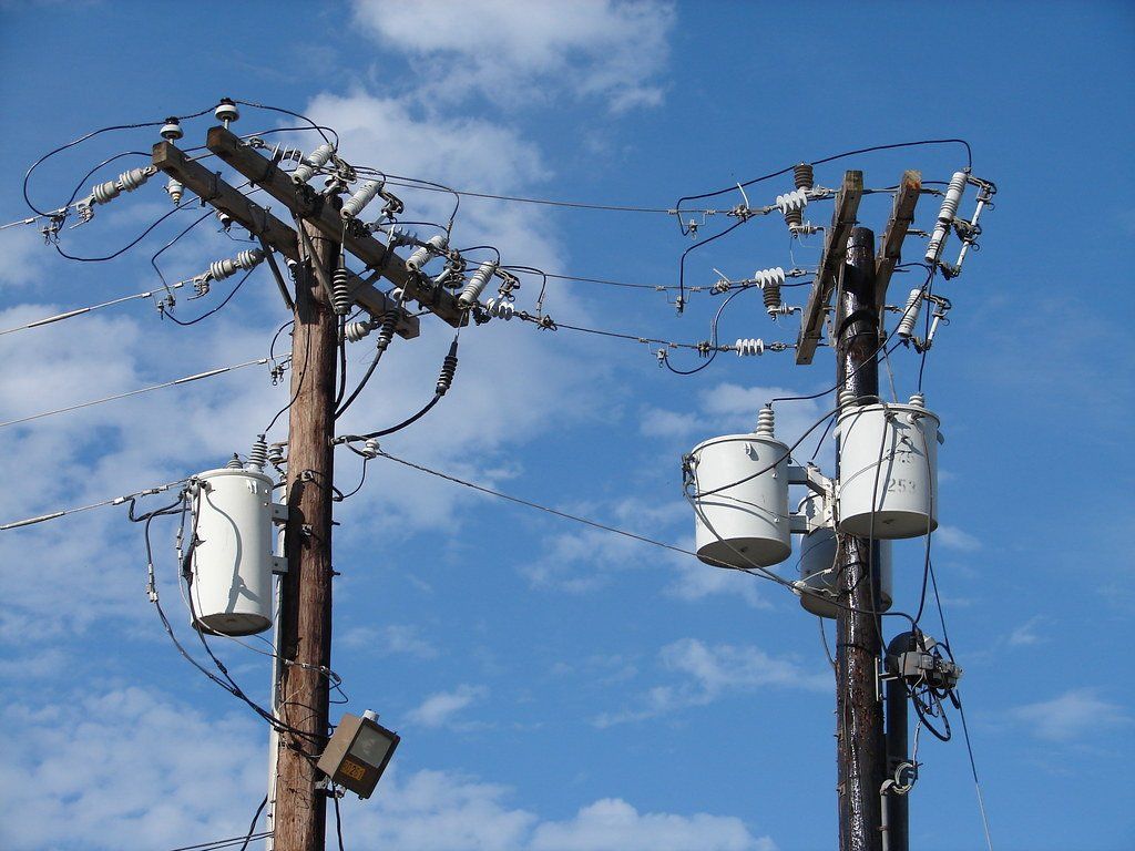 Two utility poles with transformers and electrical wires against a blue sky.