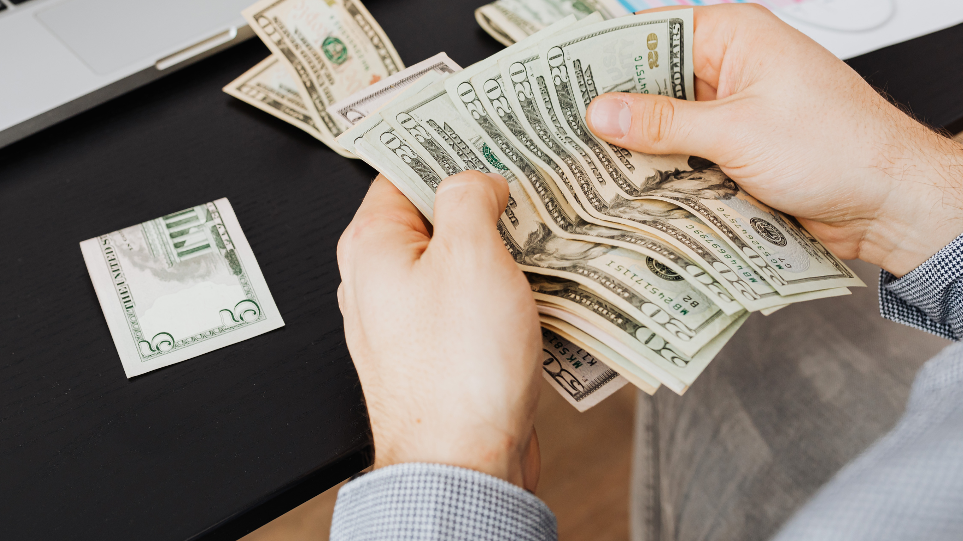 Hands counting U.S. dollar bills at a black table with more cash stacked nearby