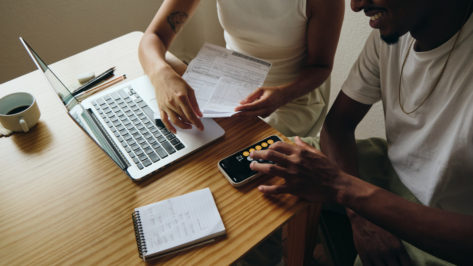 Two people collaborating at a wooden table with a laptop, papers, and a smartphone.