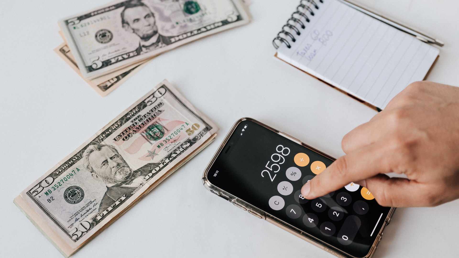 Hand tapping calculator app on smartphone beside scattered cash and a notepad on a white desk