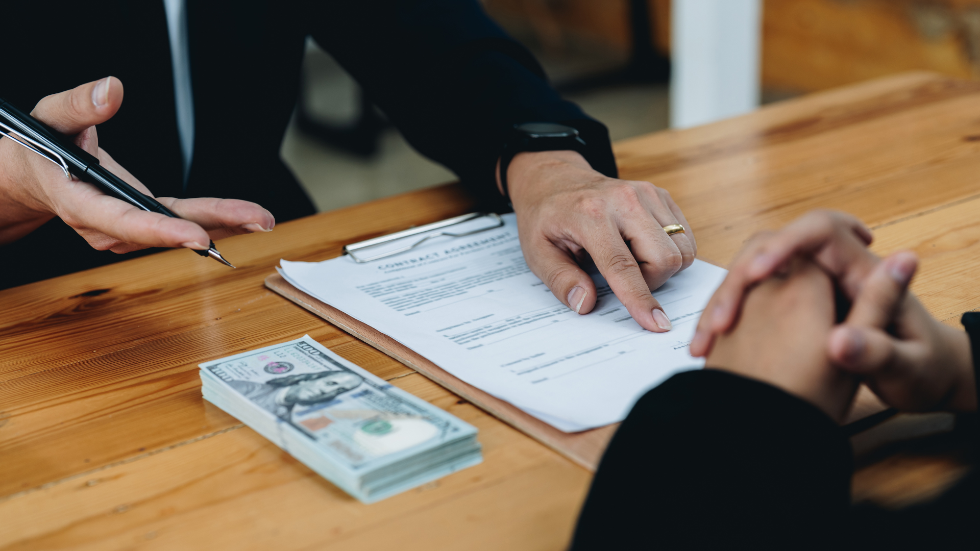 Hands signing a document on a wooden table beside a stack of cash