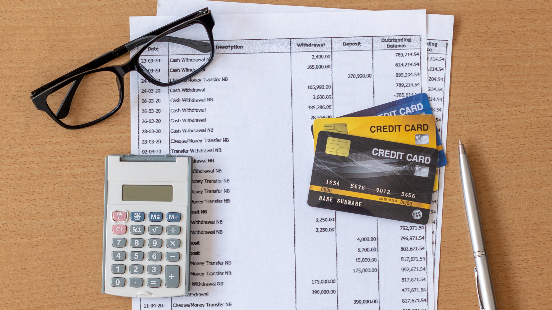 Glasses, calculator, cards, and pen on top of paperwork on a wooden desk.