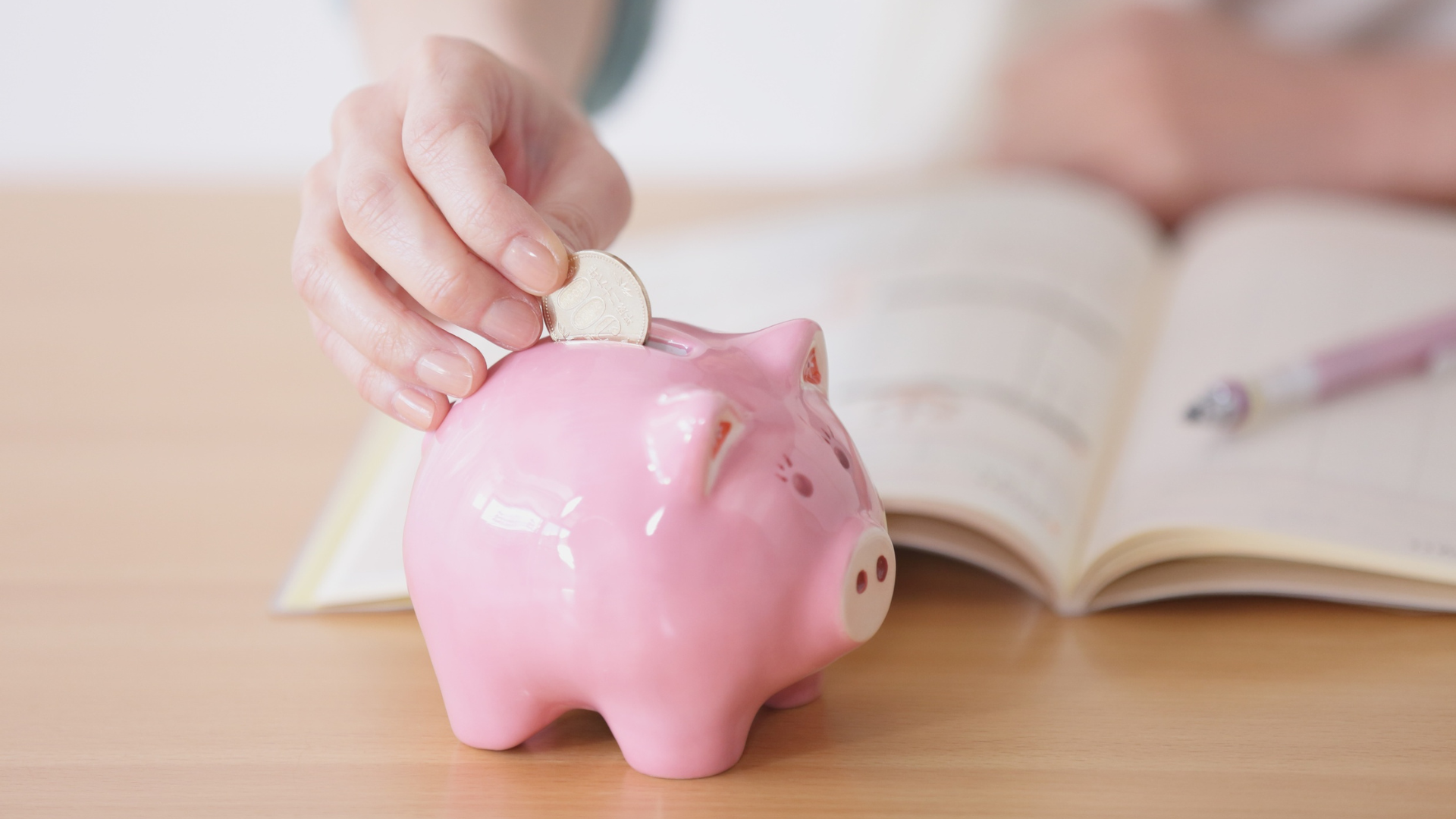 Hand placing a coin into a pink piggy bank beside an open book on a desk