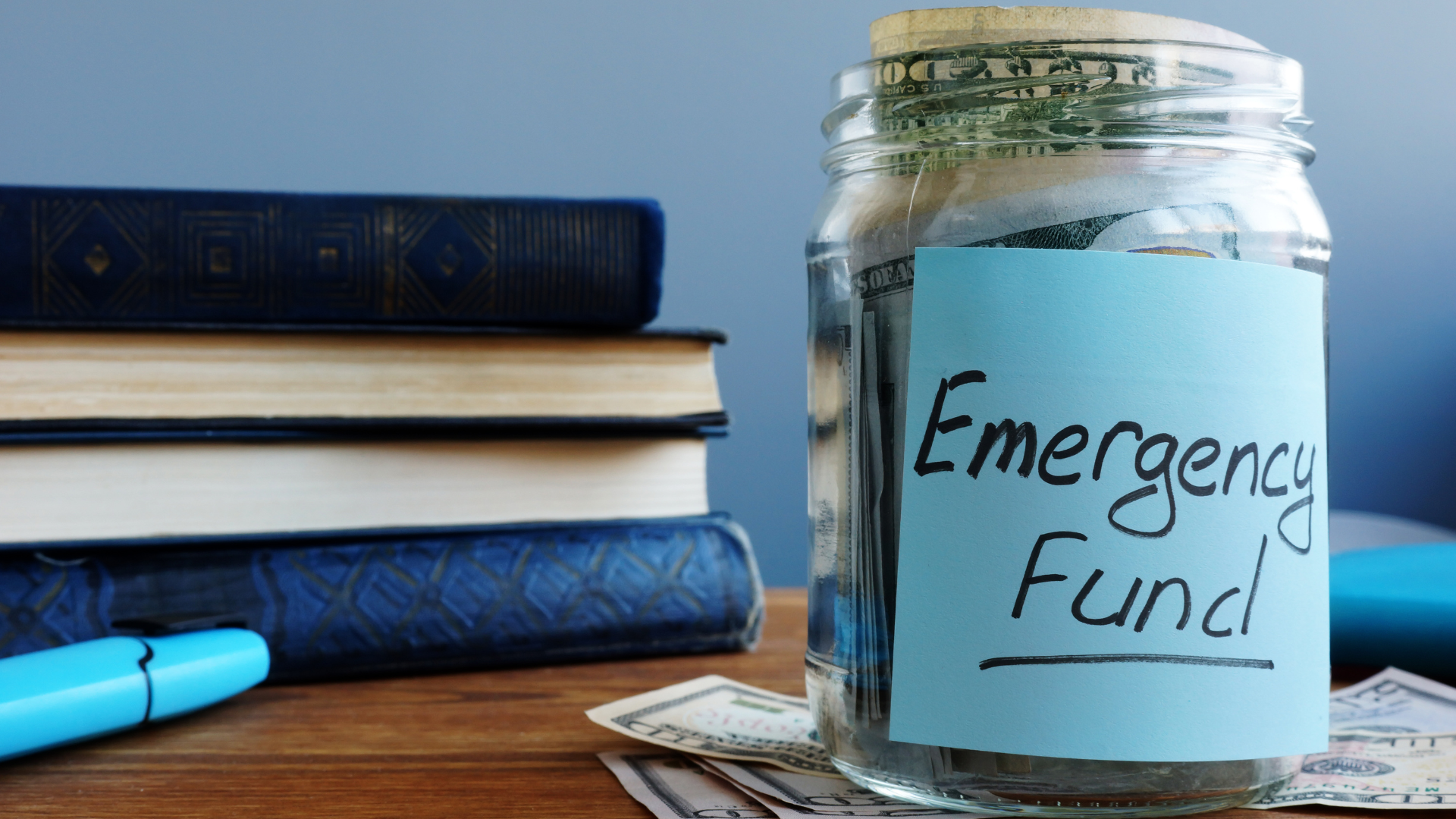 Glass jar labeled “Emergency Fund” beside stacked books and pens on a desk.