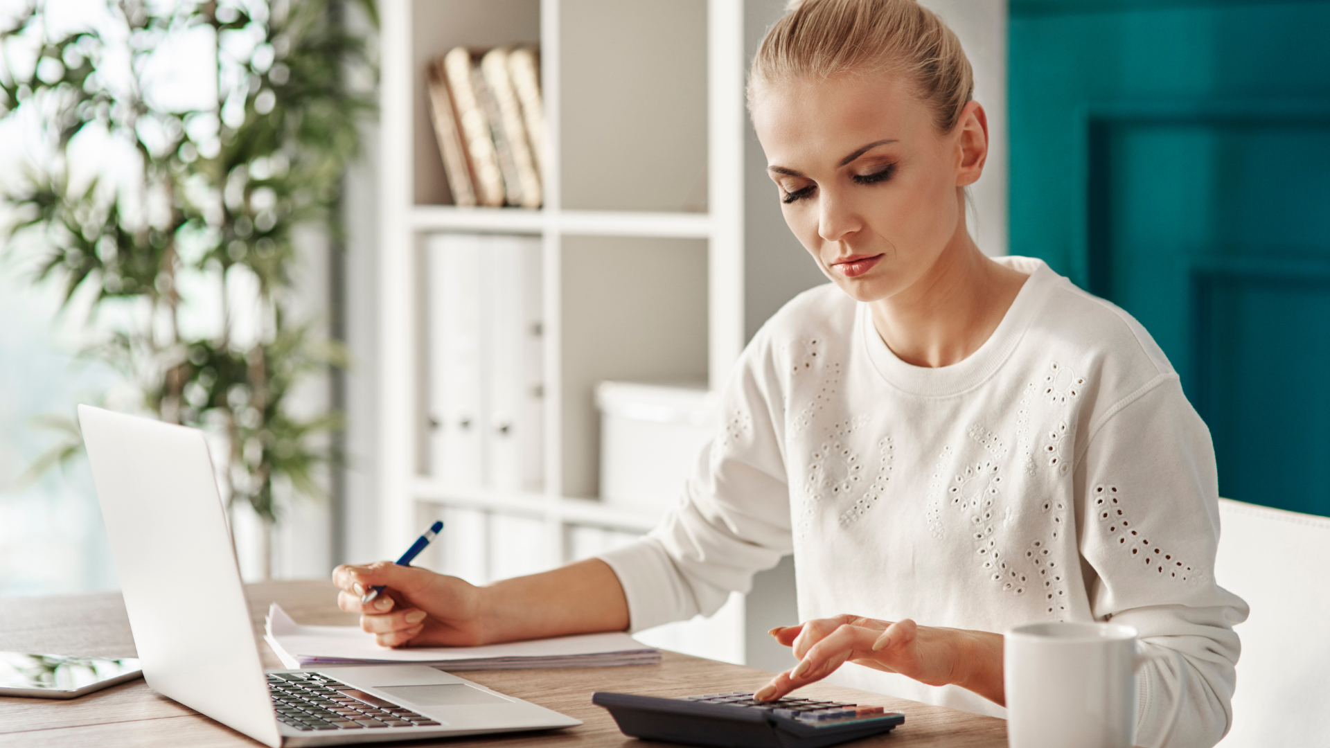 Person working at a desk with a laptop, calculator, and notebook in a bright office.