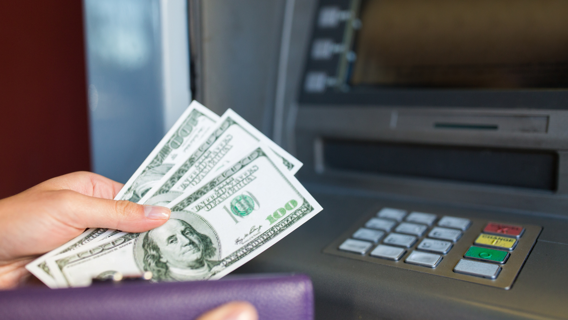 Hand holding U.S. dollar bills near an ATM keypad and cash machine slot