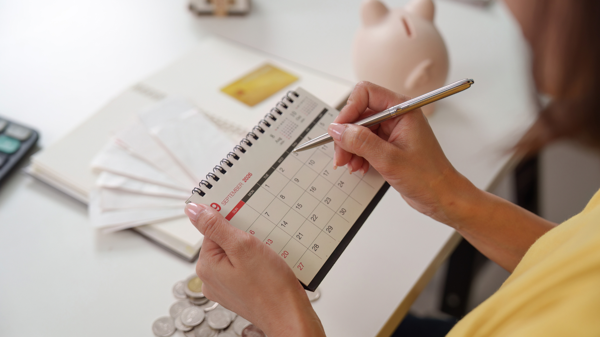 Hands writing in a planner beside stacks of bills and a piggy bank on a desk