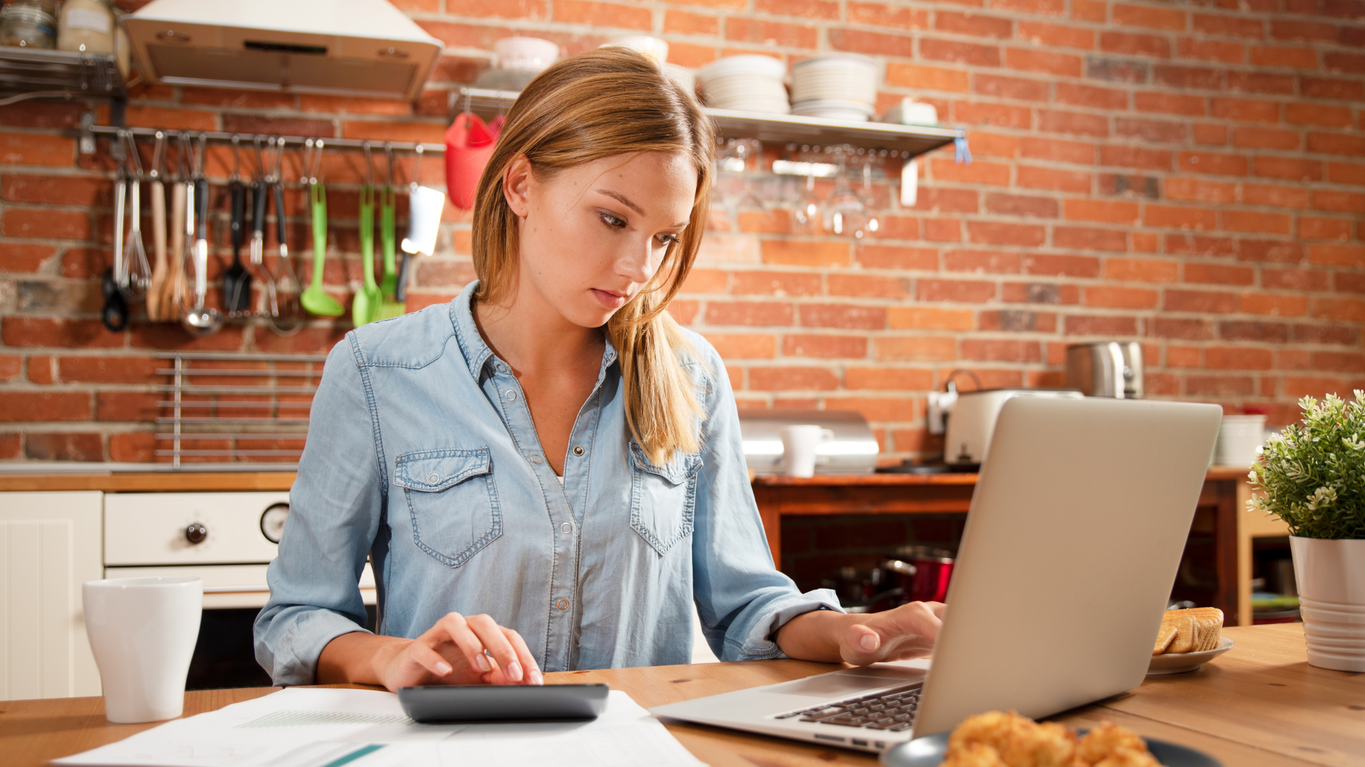 Woman working on a laptop at a kitchen table with papers and a tablet nearby, in a brick-walled kitchen