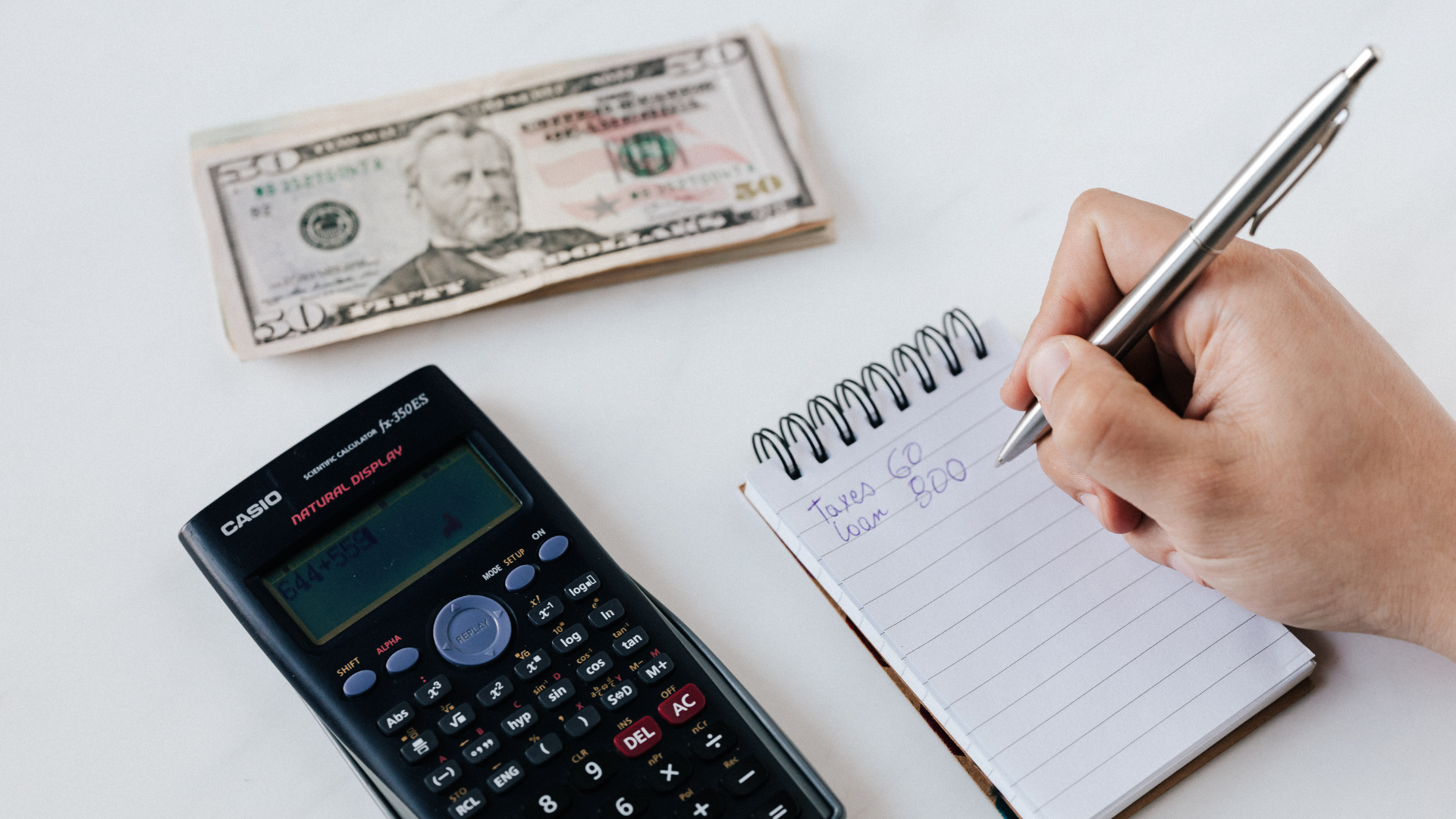 Hand writing expenses in a notebook beside a calculator and $20 bill on a white desk