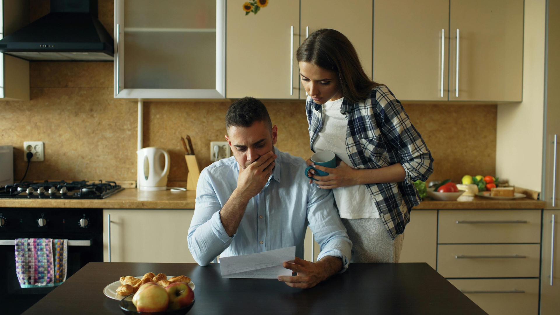 Two people in a kitchen review a document at a table, with fruit and a laptop nearby.