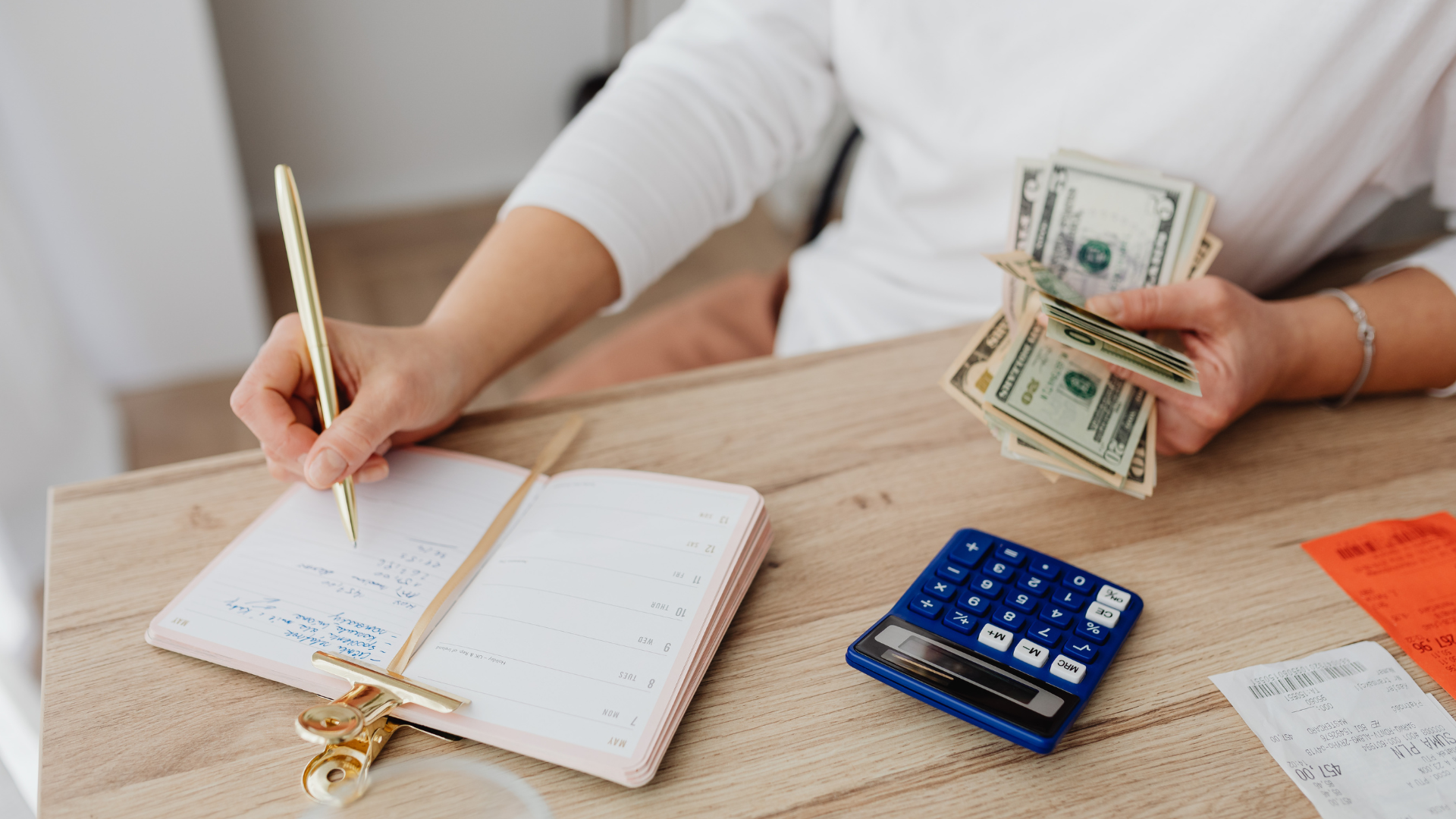Person writing in an open planner while holding cash beside a blue calculator on a table