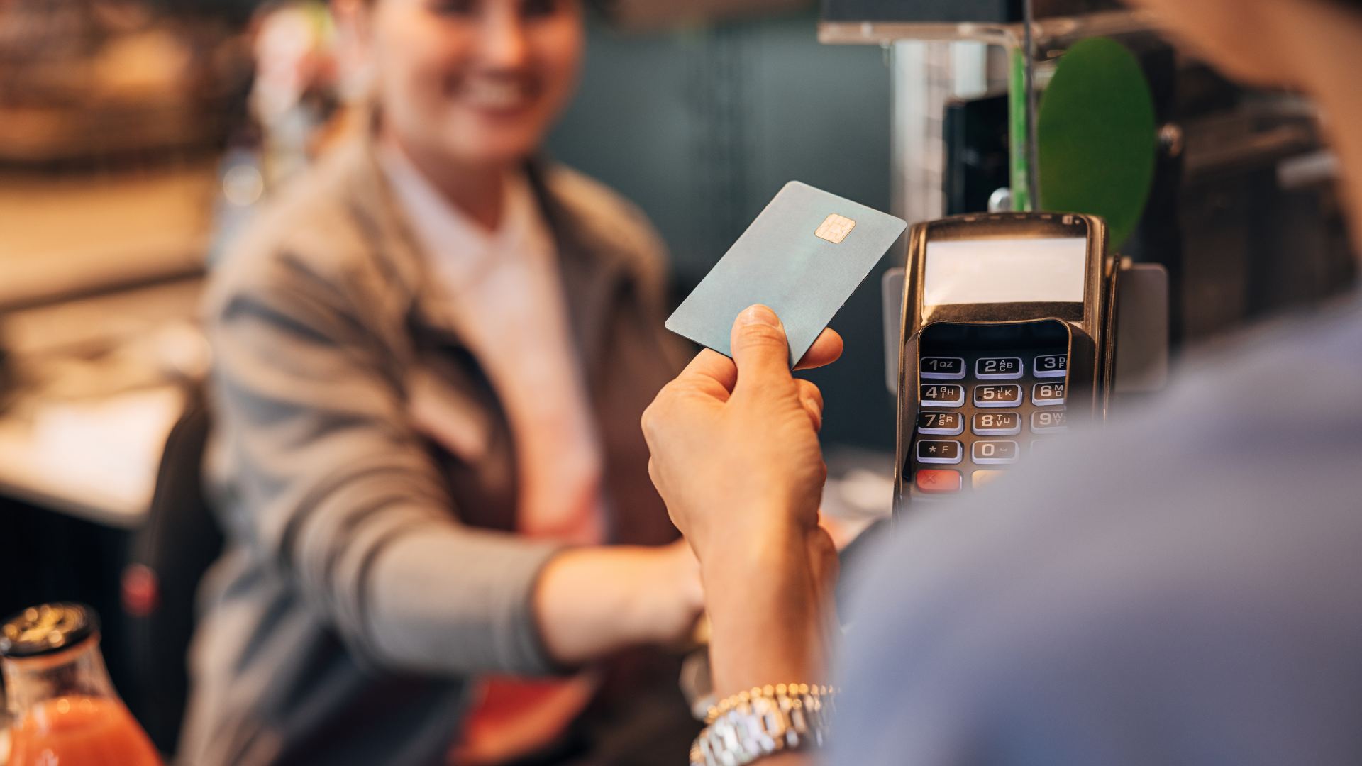 Person tapping card on payment terminal at a checkout counter, with a cashier blurred in the background
