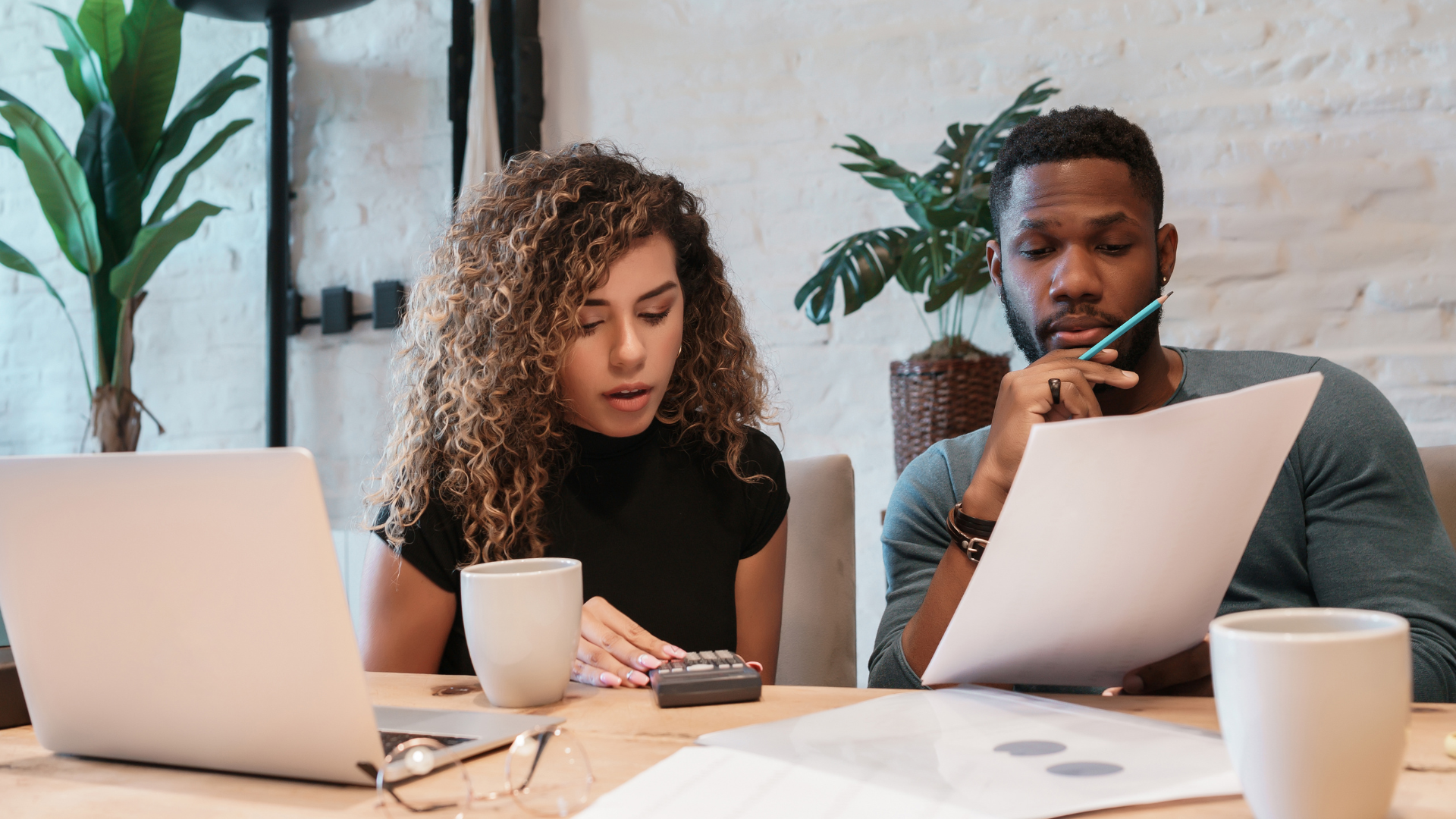 Two coworkers reviewing documents at a desk with laptops, coffee mugs, and indoor plants