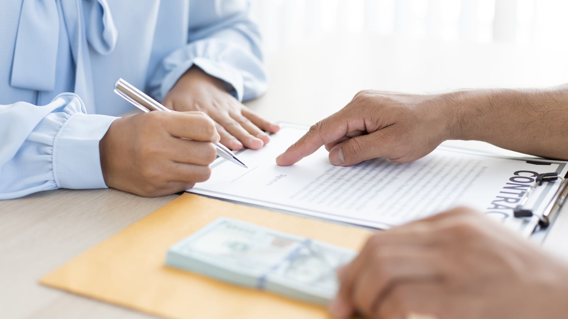Hands signing a document on a clipboard beside a stack of cash on a desk