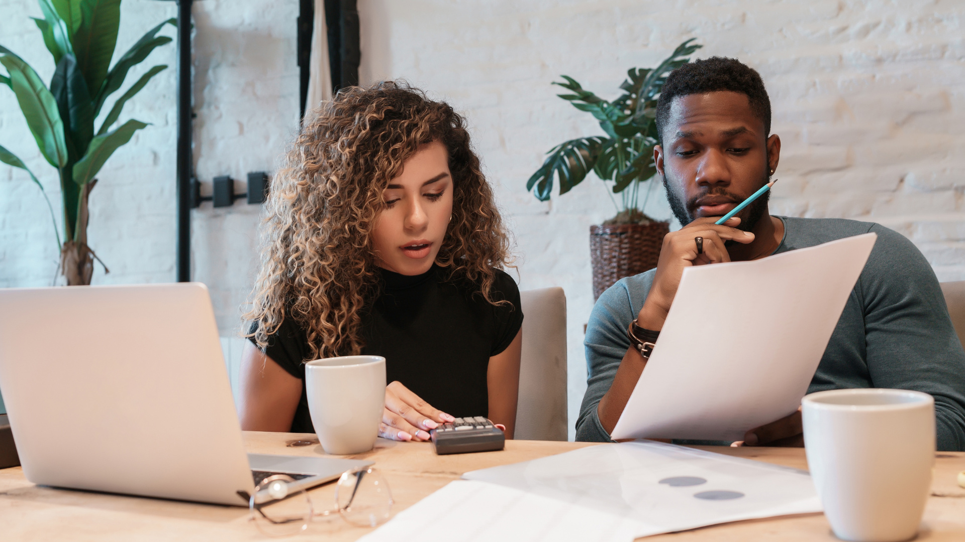 Two people working at a table with laptops and papers in a bright office
