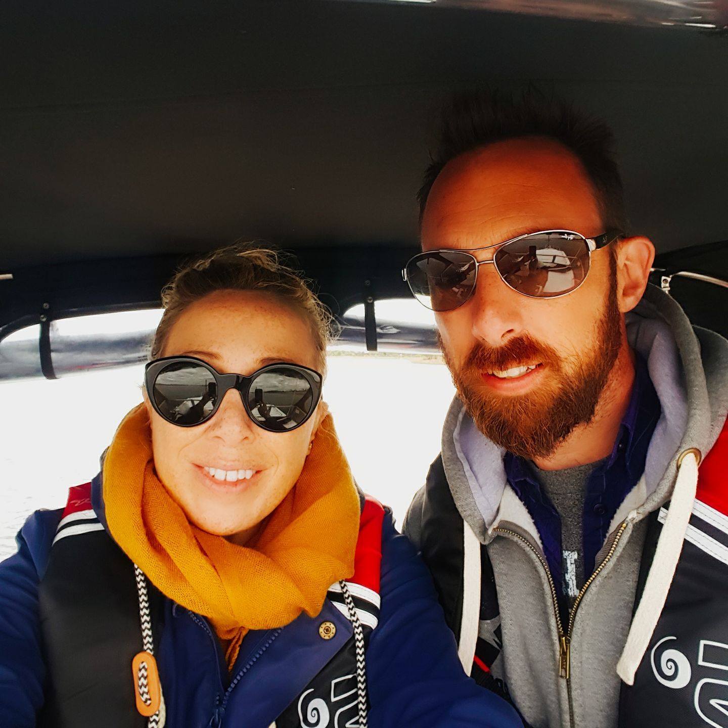 Two people in sunglasses on a boat; woman with a yellow scarf and man with a beard smile at the camera.