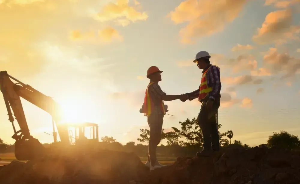 Two construction workers are shaking hands at a construction site at sunset.