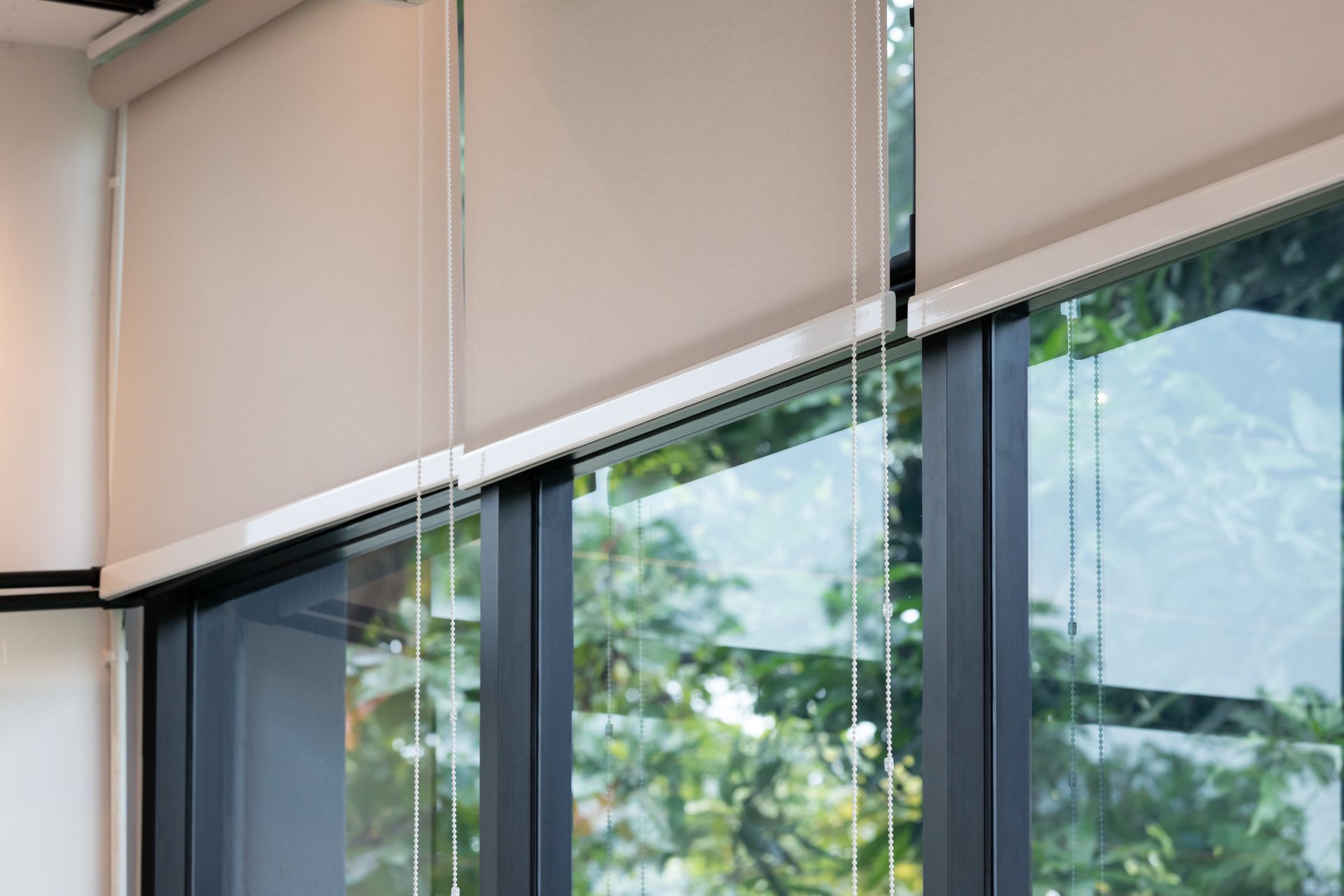 Beige roller shades partially covering a window with a view of green trees.