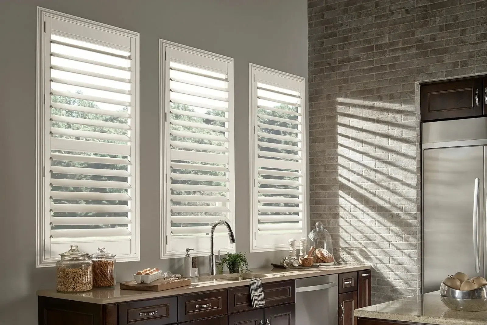 Kitchen with three white window shutters, dark cabinetry, and exposed brick wall. Sunlight creates shadows.