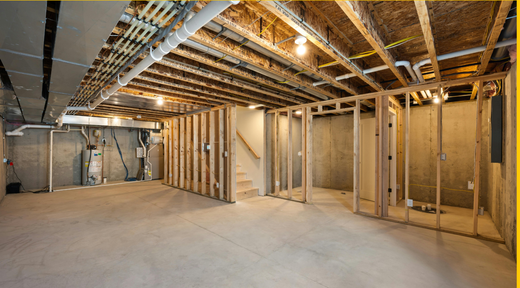 Unfinished basement with concrete floor, wooden framing, exposed pipes and wiring, and concrete walls. Framing a staircase is visible.