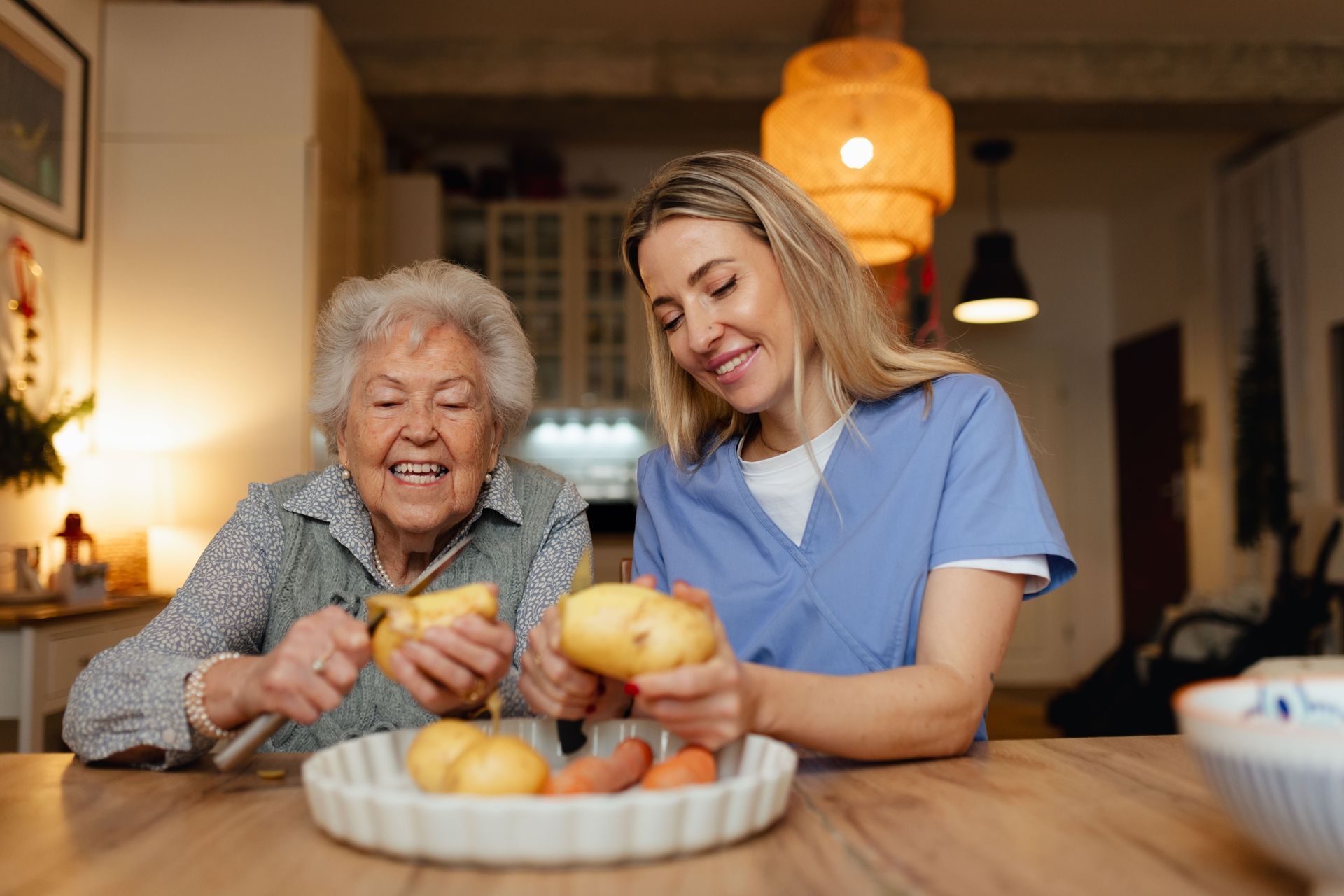 Woman and elderly person peeling potatoes together at a wooden table; both smiling.
