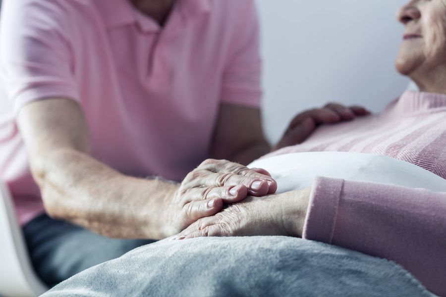 Man in pink shirt holding hands with an elderly woman in a light pink sweater; indoors, showing care and comfort.