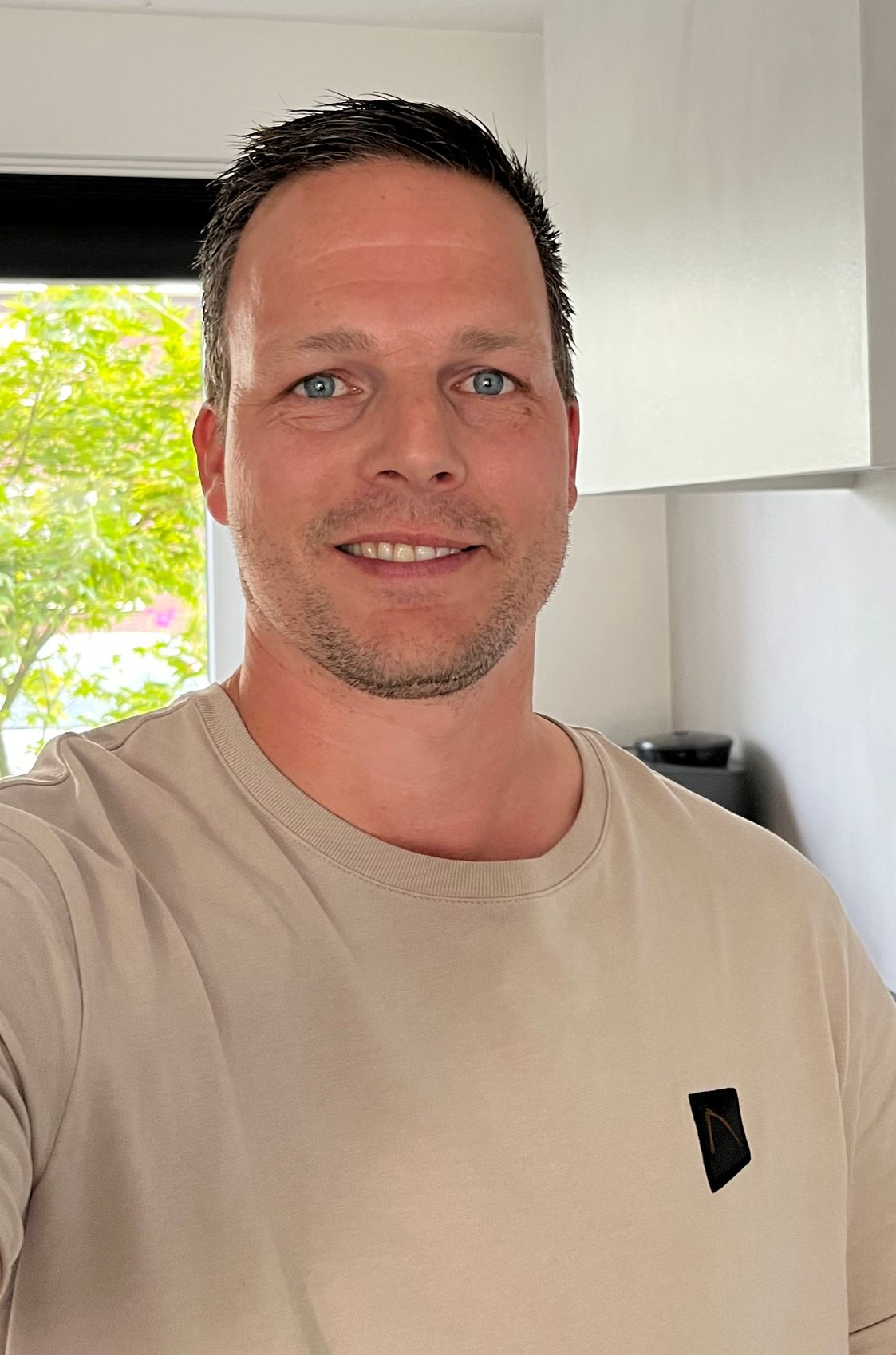 Een man met kort bruin haar glimlacht, gekleed in een lichtbruin T-shirt, en staat binnen bij een raam.