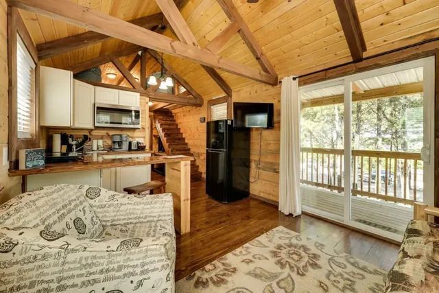 Cozy cabin interior: wood walls, kitchen, stairs, sofa, sliding glass door to balcony with trees.