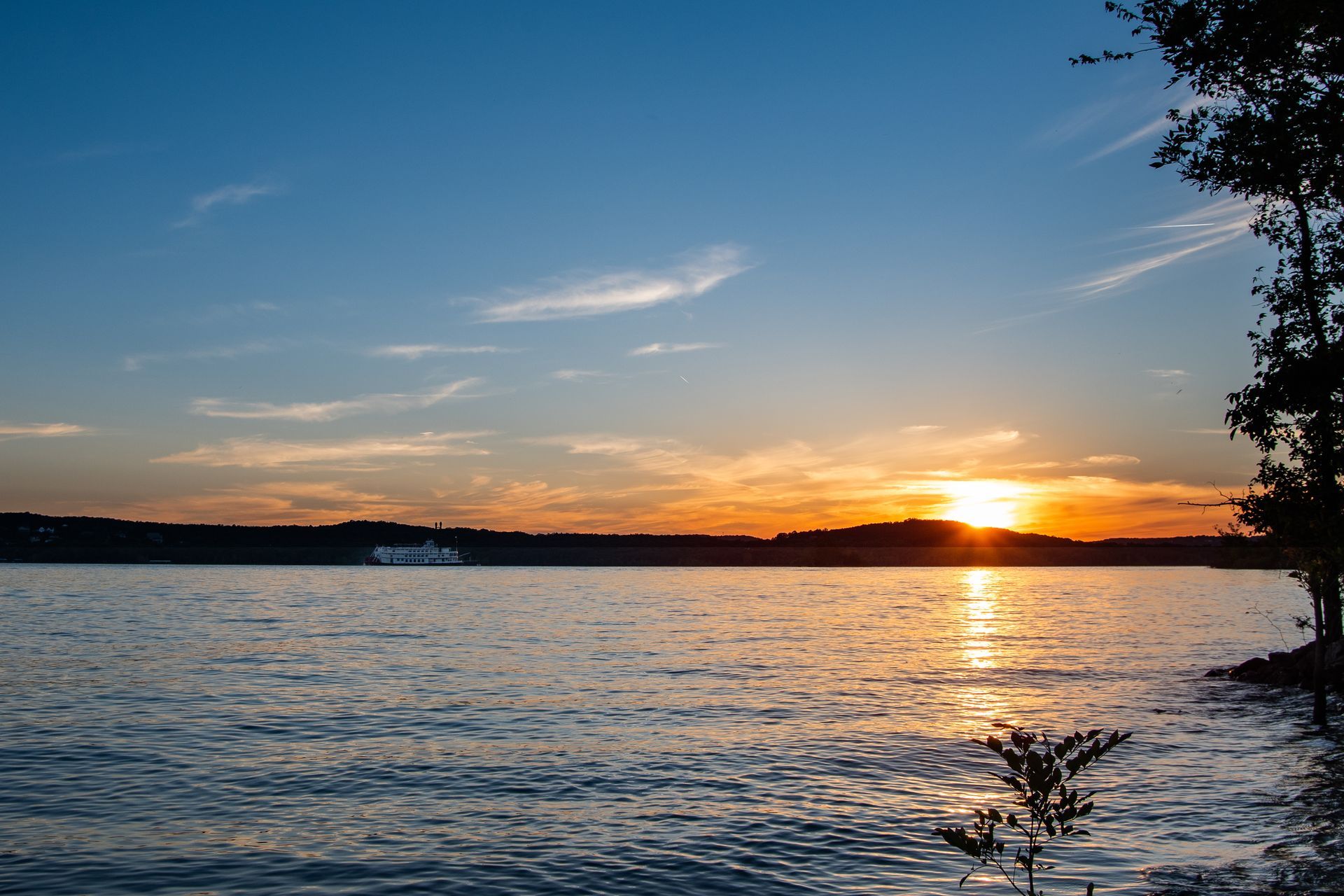 Sunset over calm water, orange and gold reflecting; tree silhouette on right, distant shoreline.