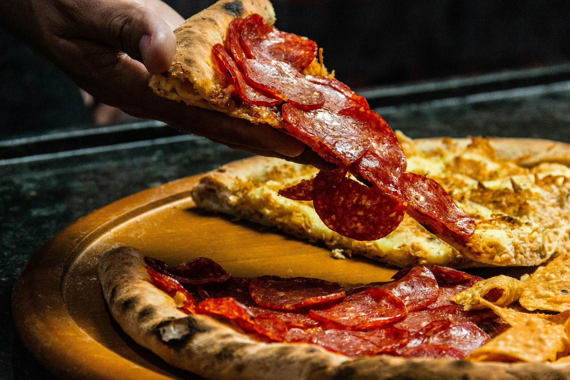 Hand holding a pepperoni pizza slice above a wooden pizza board with a pizza.