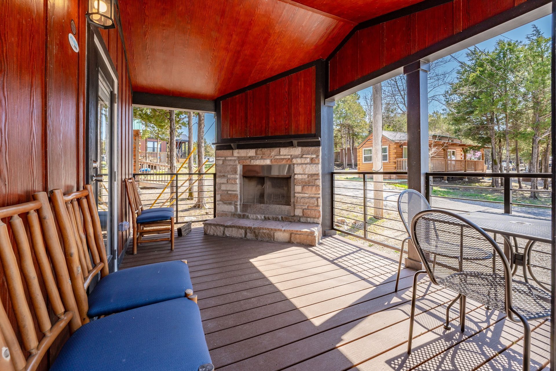 Covered porch with fireplace, seating, and view of other cabins. Brown wood, blue cushions, sunny.