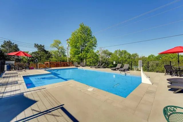 Swimming pool surrounded by a concrete patio, with lounge chairs and red umbrellas. Bright blue sky overhead.