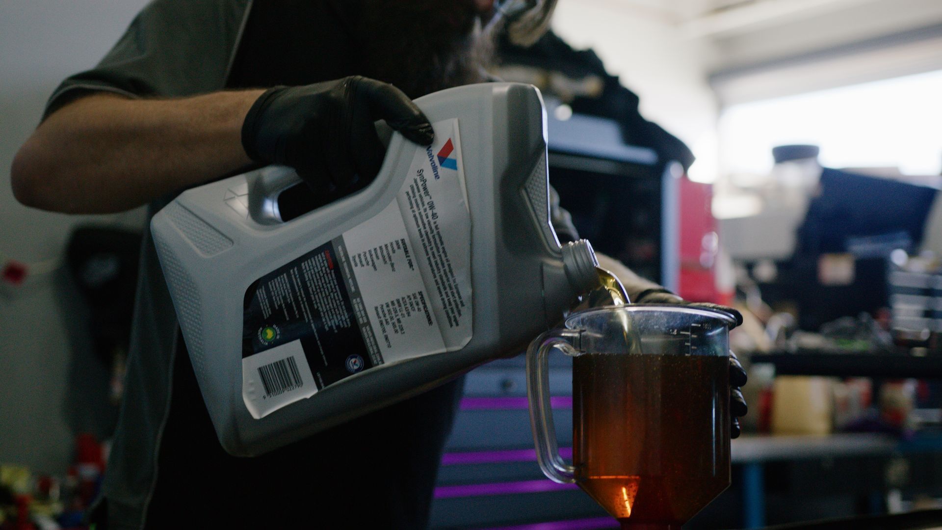 Gloved worker pours amber liquid from a gray jug into a clear container in a workshop.