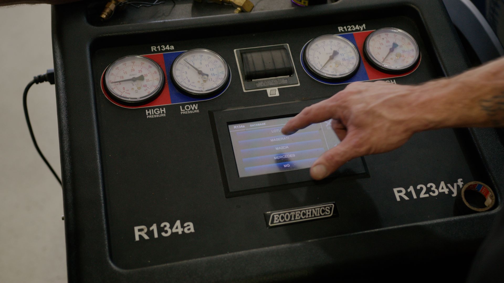 Hand pointing at touchscreen on a refrigerant gauge panel labeled R134a and R1234yf