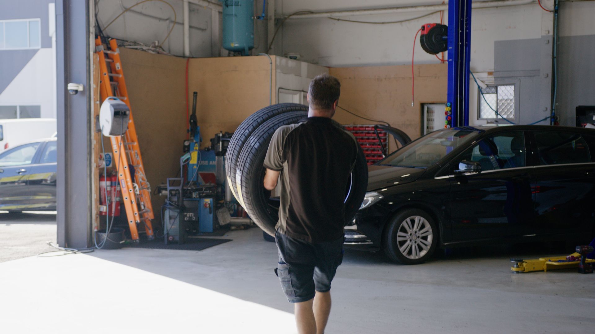 Man walking into a garage with stacked tires beside a black car