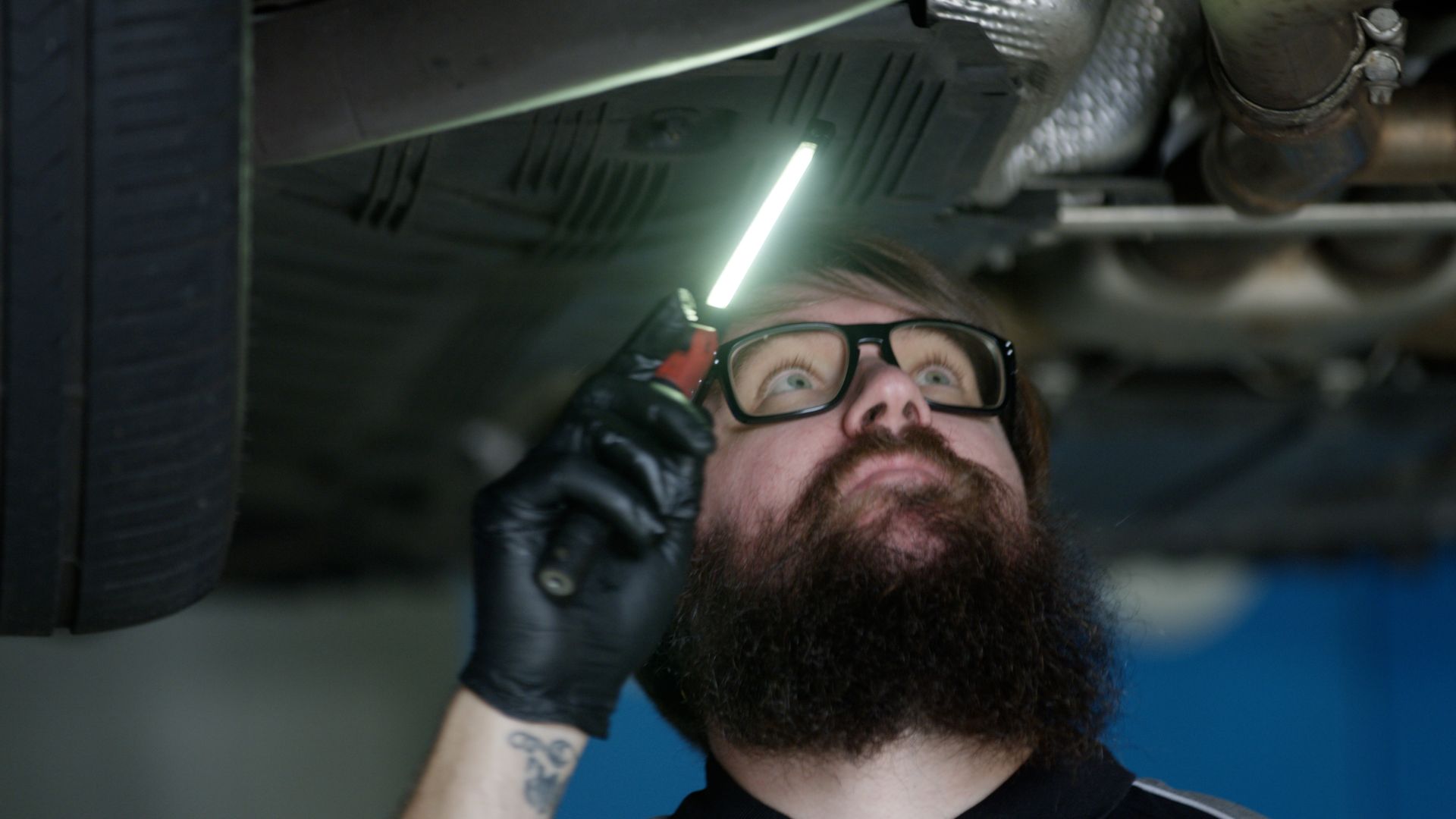 Mechanic inspecting under a vehicle with a glowing light, wearing glasses and black gloves
