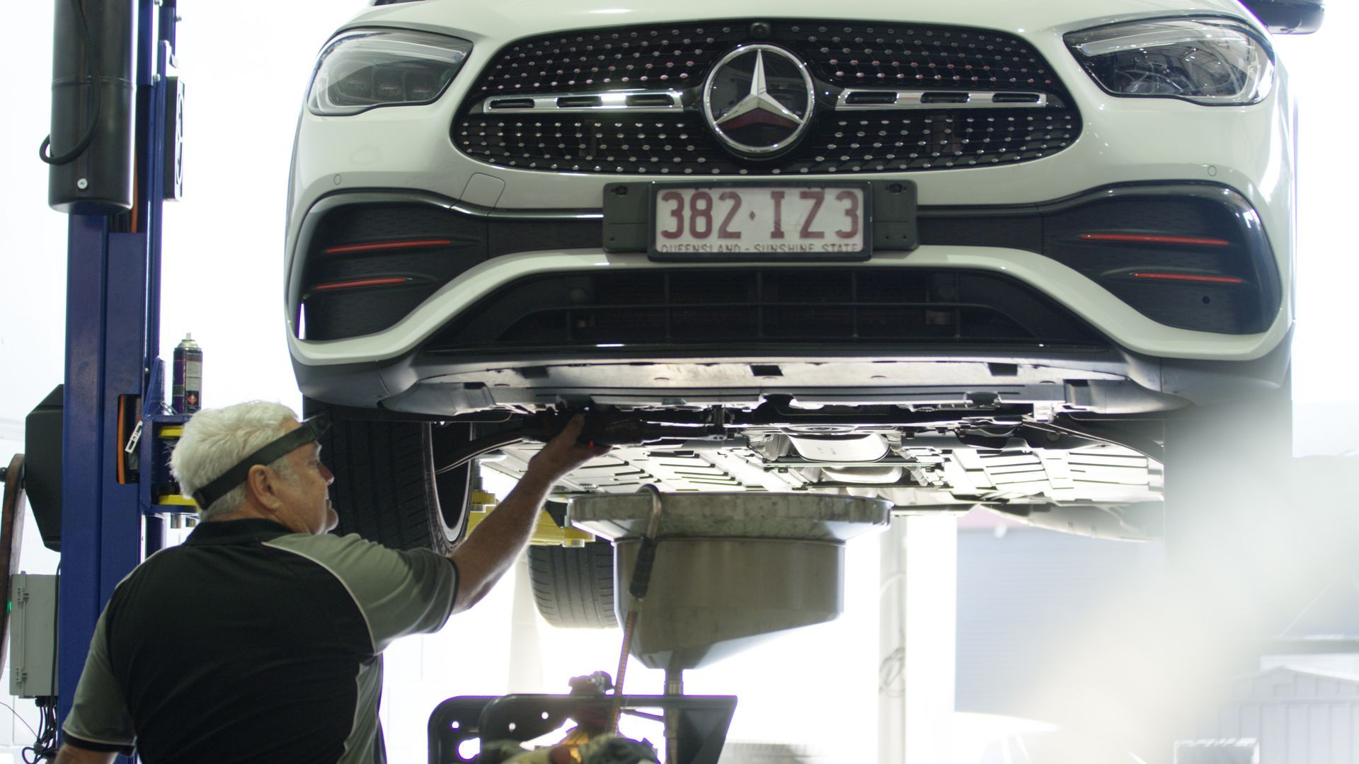 Mechanic inspecting the underside of a white Mercedes-Benz on a lift in a garage