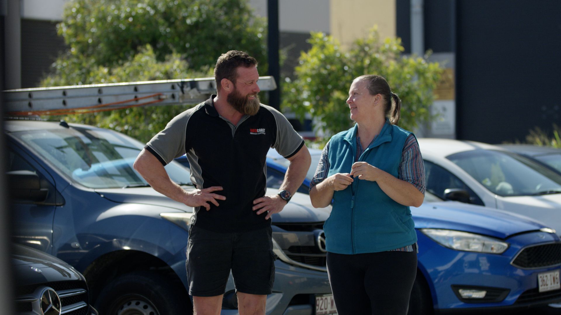 Two people talking beside parked cars in a sunny lot, one in a black shirt and the other in a blue top.
