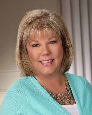 Portrait of a smiling person in a turquoise sweater and necklace against a neutral studio backdrop