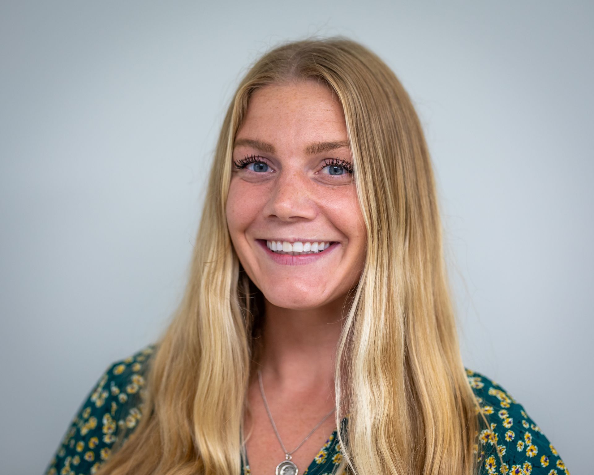 Smiling person with long blonde hair in a green patterned top against a gray background
