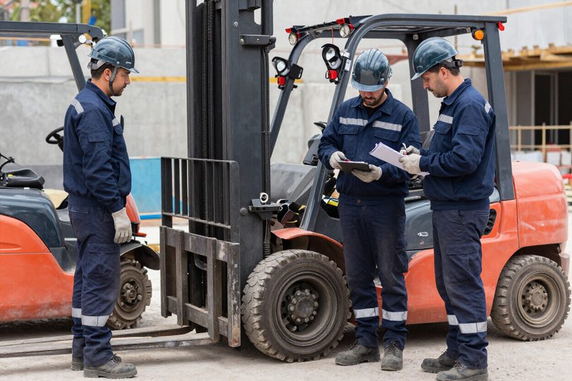 Tres trabajadores con cascos de seguridad inspeccionan una carretilla elevadora en un patio industrial.