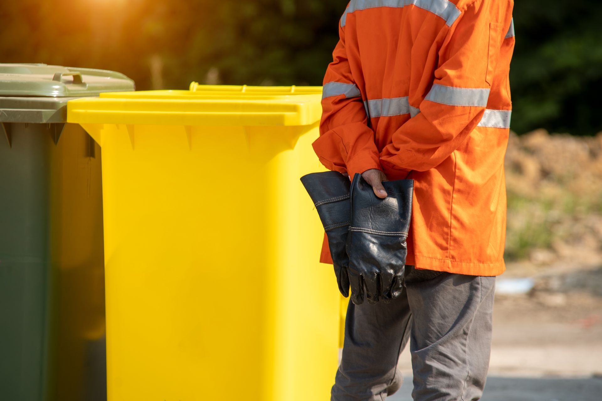 A rubbish collection worker holding gloves & dressed in an orange jacket is standing by two bins.