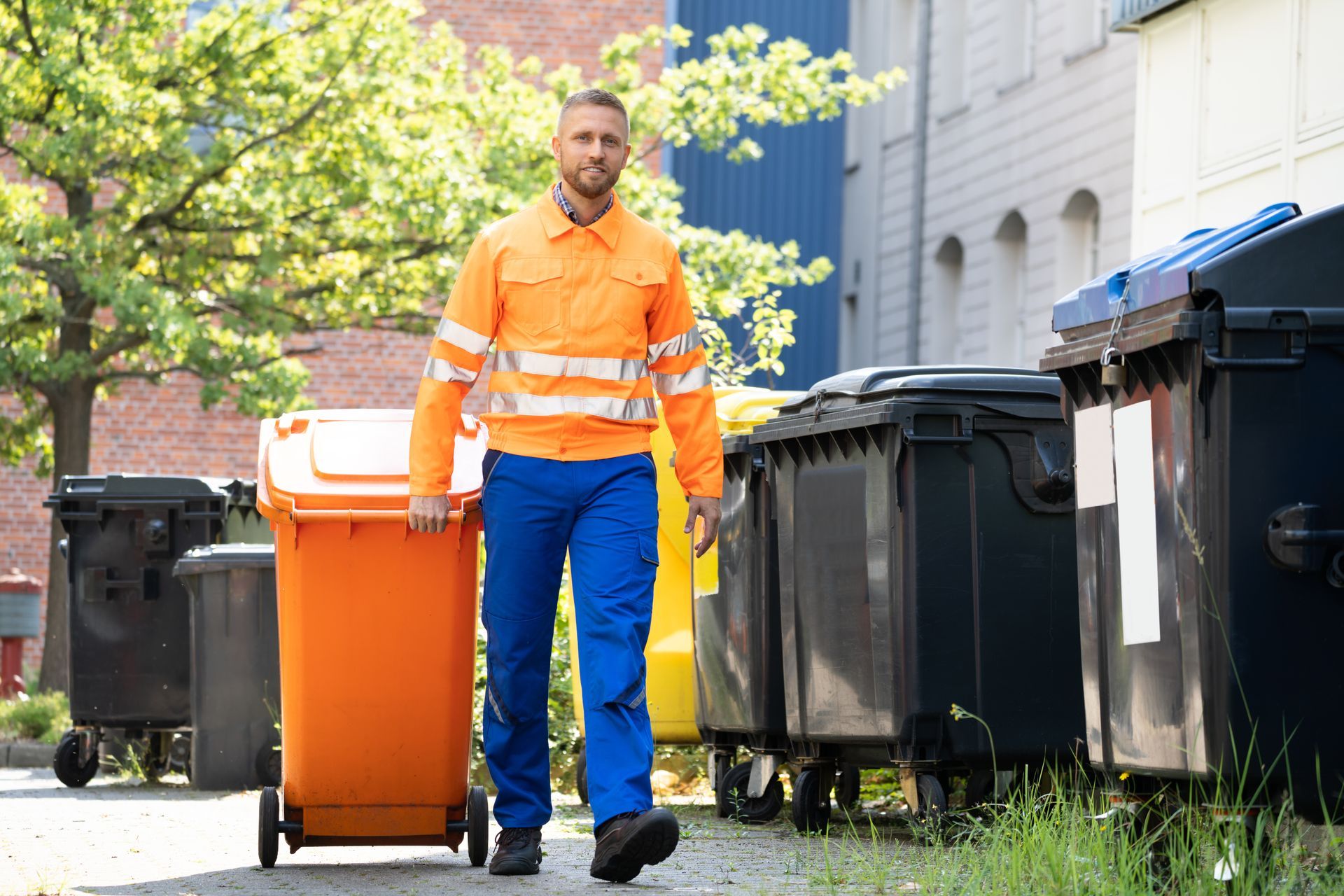 AN INDUSTRIAL WORKER WHEELING A SMALL SKIP BIN OUTDOORS NEXT TO LARGE BINS.
